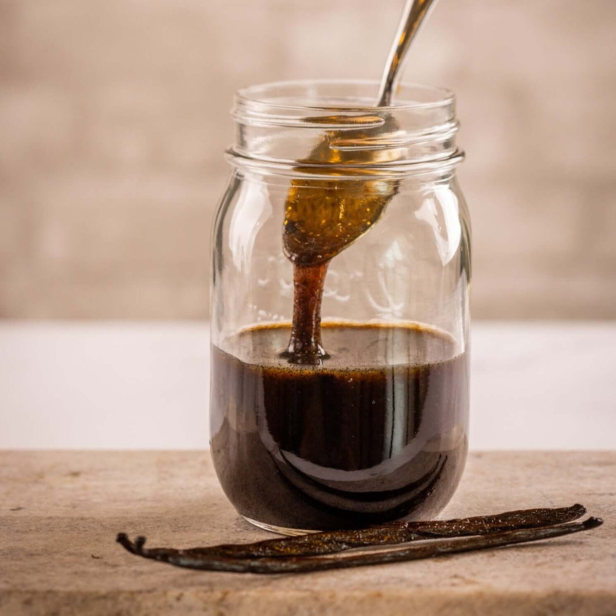 Madagascar Pompona Vanilla Beans extract in a jar with spoon and vanilla pods on the table.